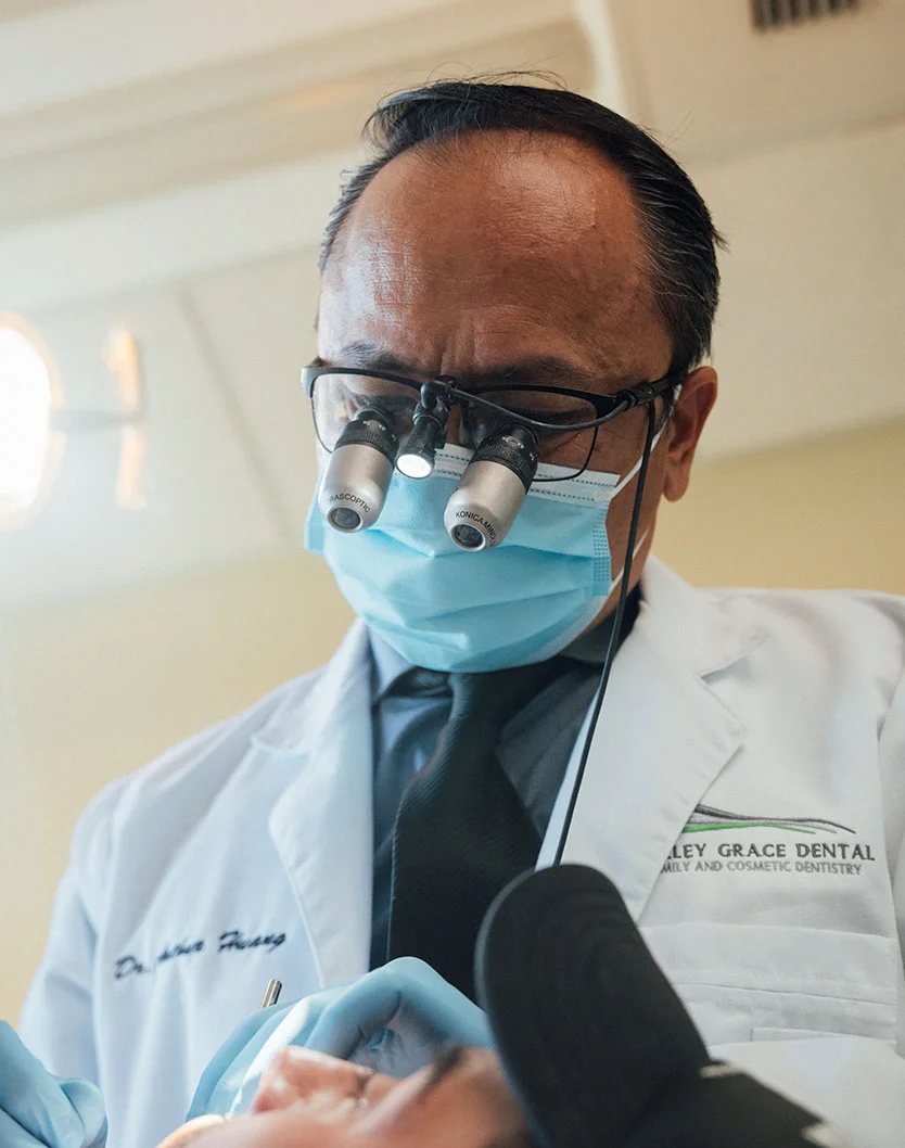 Dr. Huang, wearing a mask and scrubs, is holding a book in a healthcare setting. The presence of medical equipment suggests they are in a medical or dental clinic, possibly involved in healthcare practices or education. - Deep Cleaning in Bakersfield, CA