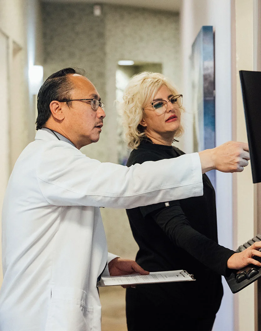 Dr. Huang and an associate, both dressed in formal clothing, are standing indoors and examining a computer screen together. The background includes a wall, and their faces are visible as they engage with the screen. - Dental Emergencies in Bakersfield, CA