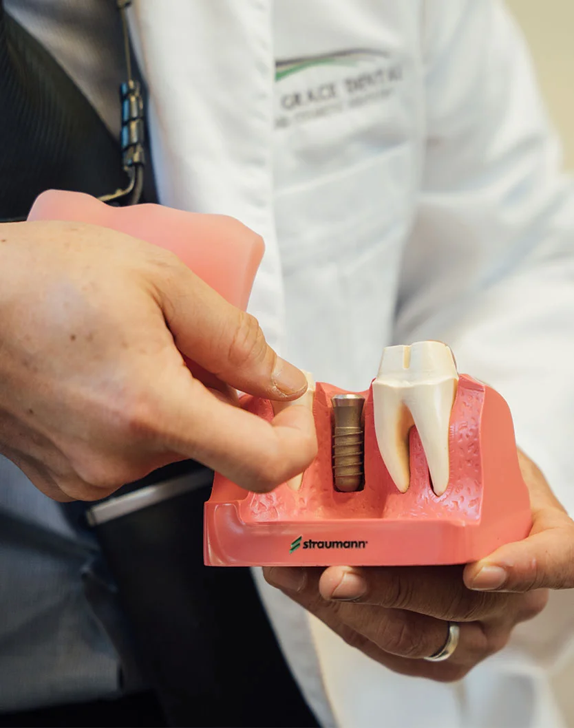 A close-up view of a dental professional carefully examining a dental model featuring a Straumann implant. The model, showcasing a reddish-pink jaw with a precisely placed implant and an abutment tooth, rests on a cream-colored base. The professional's gloved hands demonstrate a meticulous approach, highlighting the importance of precision and attention to detail in dental implant planning and treatment. - Dental Implants in Bakersfield, CA