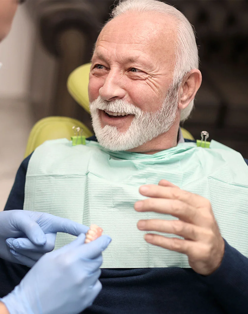 A close-up view of a senior patient with a full white beard, smiling broadly as he interacts with his dentist. The dentist presents a partial denture, and the patient's engagement and positive expression highlight the importance of personalized and compassionate dental care. - Dentures in Bakersfield, CA