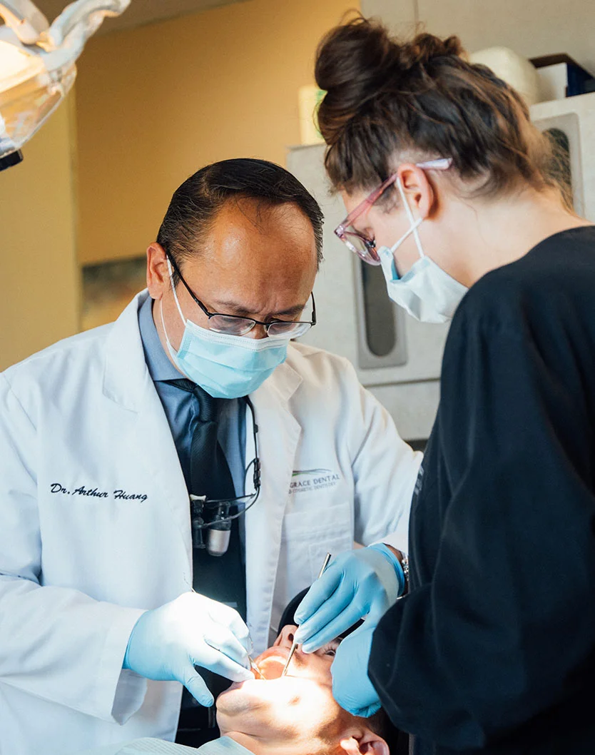 A close-up view captures a dental procedure in progress. Dr. Huang, wearing a white lab coat, a blue surgical mask, and protective eyewear, meticulously works on a patient, utilizing specialized dental instruments. The presence of a dental assistant, wearing a dark scrub top and assisting with the procedure, highlights the collaborative approach to patient care. - Exams and Cleanings in Bakersfield, CA