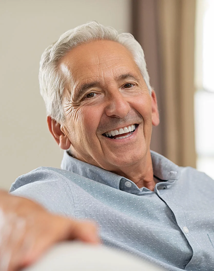 A smiling senior man with wrinkles points while indoors. He is wearing a fabric shirt, and the setting suggests an indoor environment. - Implant Overdentures in Bakersfield, CA