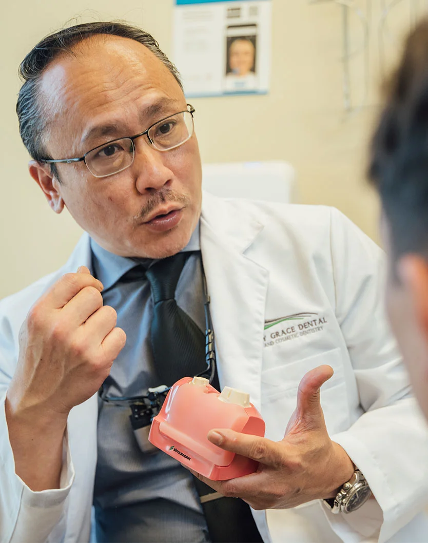 Dr. Huang, a skilled dentist with a focus on patient care, attentively examines a pink dental model, likely explaining a treatment plan or concept to a patient. His focused expression and the presence of a patient in the background convey a sense of personalized attention and commitment to providing high-quality dental care. - Laser Gum Contouring in Bakersfield, CA