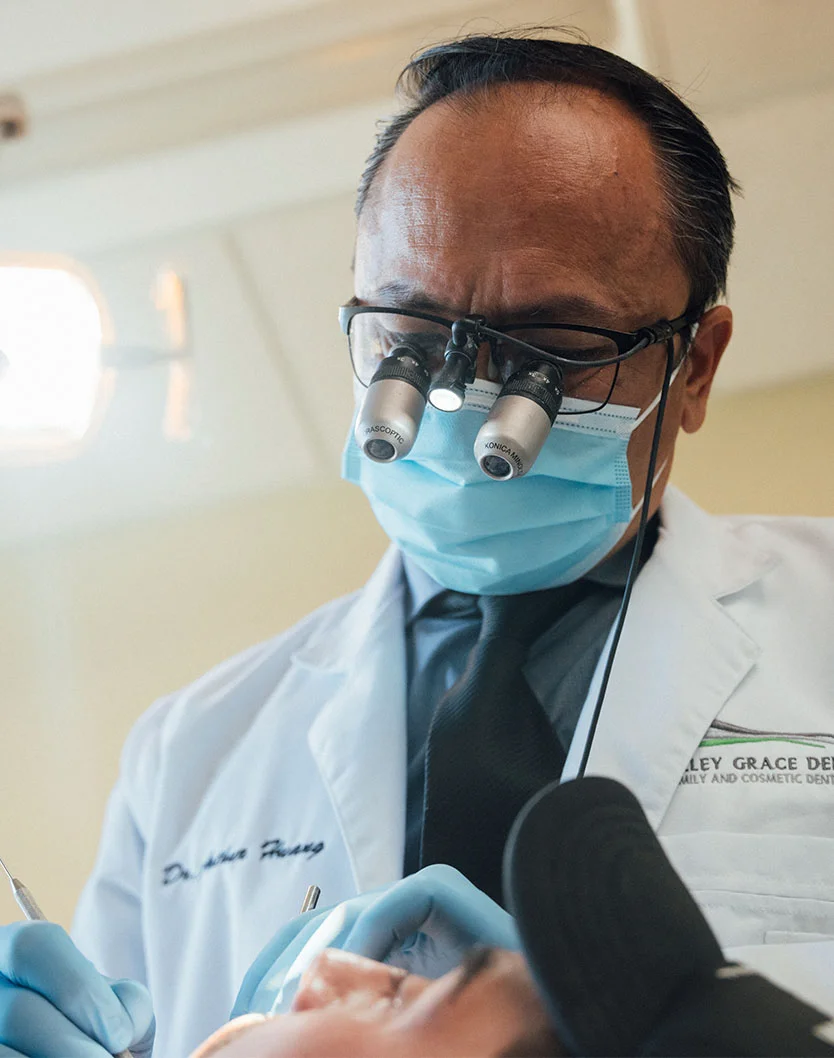 A close-up view of a dental examination in progress. Dr. Huang, wearing a white lab coat, blue surgical gloves, and magnification lenses with an attached light source, carefully examines the patient's mouth. The patient, wearing a dental bib, reclines while Dr. Huang utilizes specialized tools to ensure thorough and precise care.- Root Canal in Bakersfield, CA