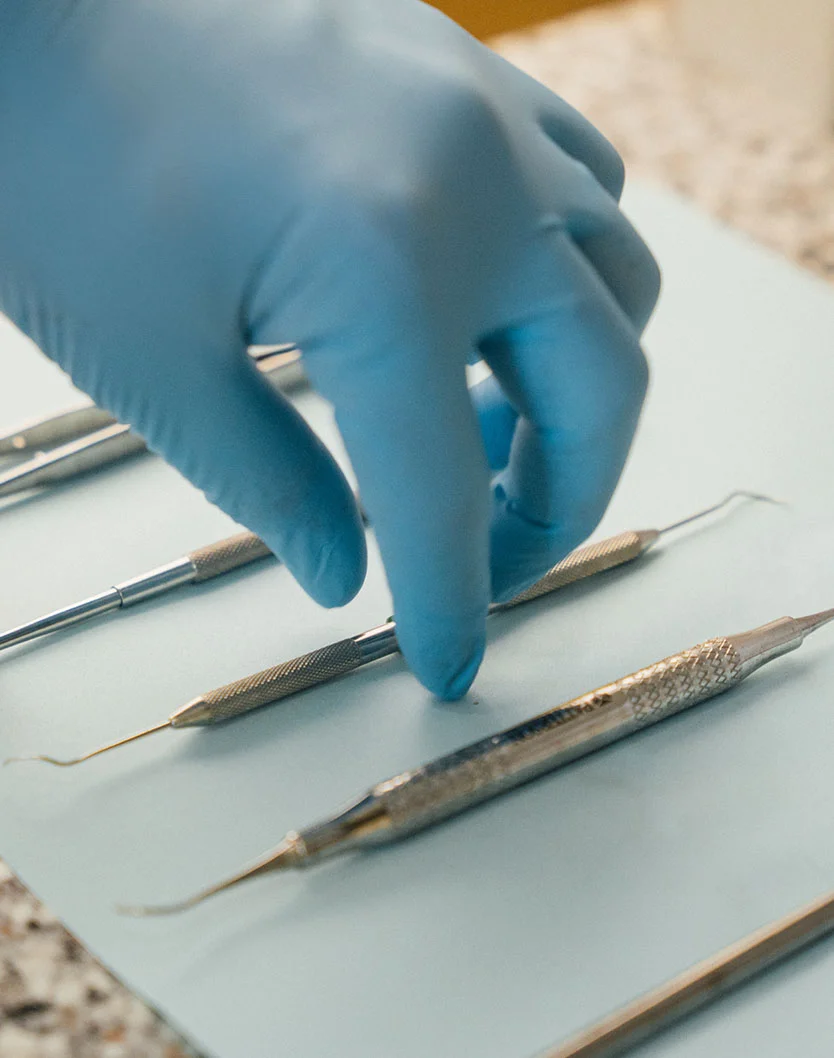 A high-angle close-up view of a dentist's hand in a blue nitrile glove reaching towards a tray of various dental instruments. The instruments are arranged on a light-blue surface within a sterile dental setting. - Tooth Extractions in Bakersfield, CA