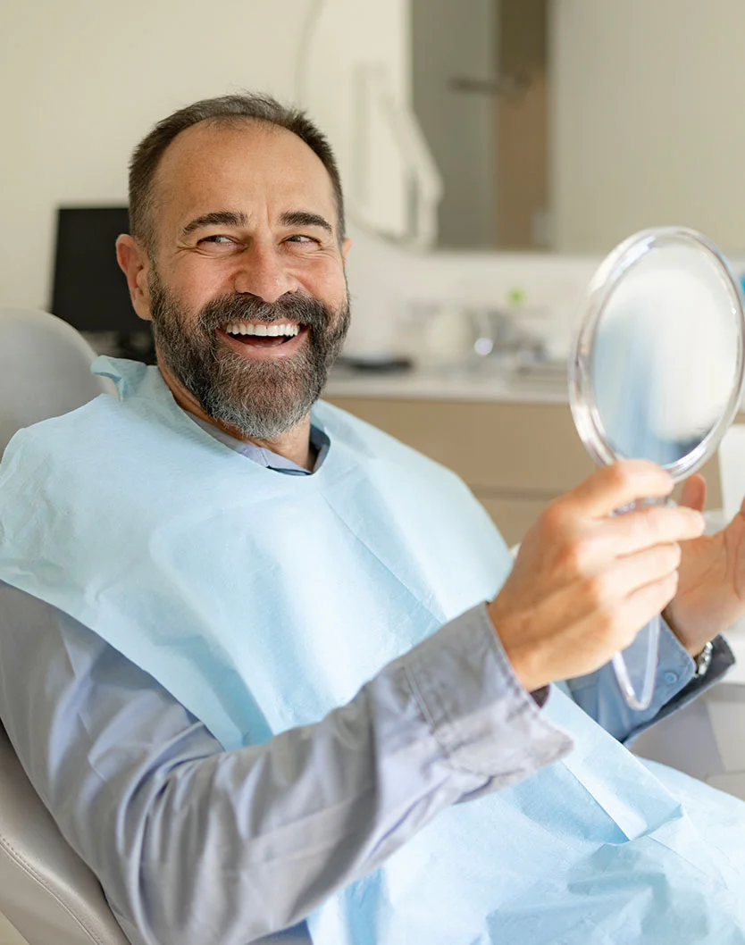 Mature man with grey beard, holding a small mirror and smiling while sitting in a dentist chair - All on 4 in Bakersfield, CA