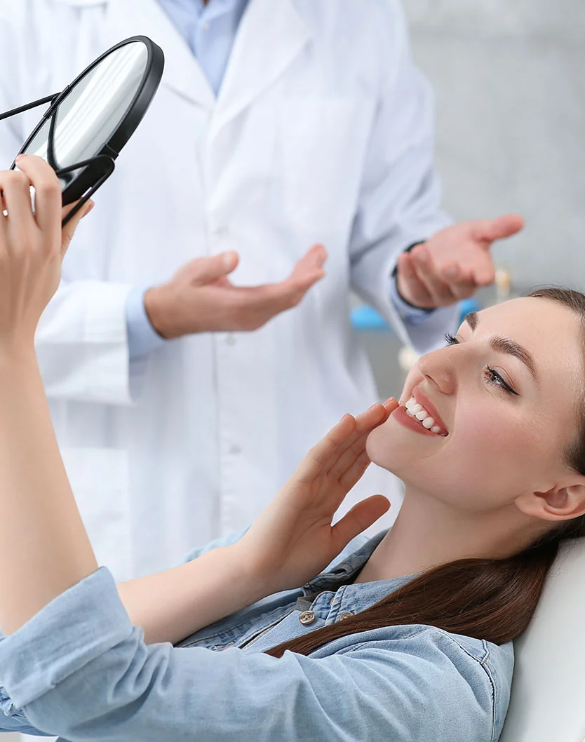 A female patient smiling while examining her teeth in a handheld mirror as her dentist explains the result - Dental Crowns in Bakersfield, CA