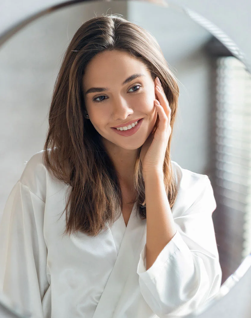 Woman with brown hair, wearing a white robe, smiling in a mirror while touching her cheek - TMJ Treatment in Bakersfield, CA